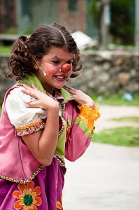 Foto de garota sorridente com o rosto pintado e nariz de palhaço, colete pink e verde sobre blusa branca de mangas bufantes e saia cor de maravilha com flores aplicadas. 