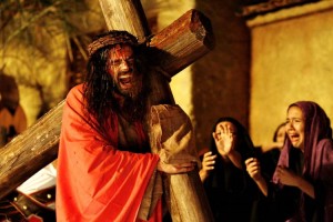 Foto colorida de Jesus, interpretado por Thiago Lacerda, carregando a cruz com manto vermelho nas costas e coroa de espinhos na cabeça. Ao seu lado, mulheres choram.