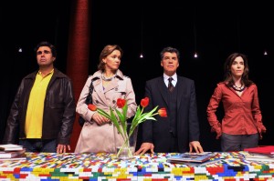 Foto do elenco em pé, atrás de mesa retangular coberta por peças coloridas de Lego e jarro com tulipas vermelhas. Da esquerda para a direita, Orã Figueiredo, Julia Lemmertz, Paulo Betti e Deborah Evelyn.