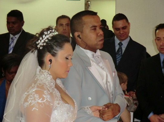 Fotografia colorida, da cintura para cima, de Rafael e Roberta de braços dados na cerimônia de casamento, os dois com fones de ouvido intra-auriculares. Ao fundo três padrinhos e o irmão da noiva.