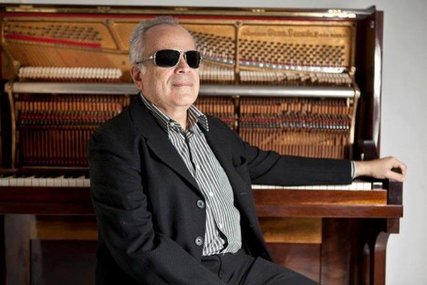 Fotografia colorida, em plano médio, de Sérgio Sá, homem de cabelos grisalhos, usando óculos escuros, terno cinza chumbo sobre camisa listrada de preto e branco. Ele está sentado com o braço apoiado sobre as teclas de um piano com cordas e martelos à mostra.