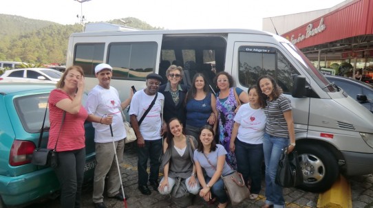 Fotografia colorida da Caravana da Audiodescrição ao lado da van prata com a porta aberta, no restaurante Frango Assado. Da esquerda para a direita: Fátima, José Vicente, Sr. Fernando, Lívia, Rosilene, Teodora, Marilene e Maísa (em pé); Patrícia e Maíra abaixadas na frente do grupo.