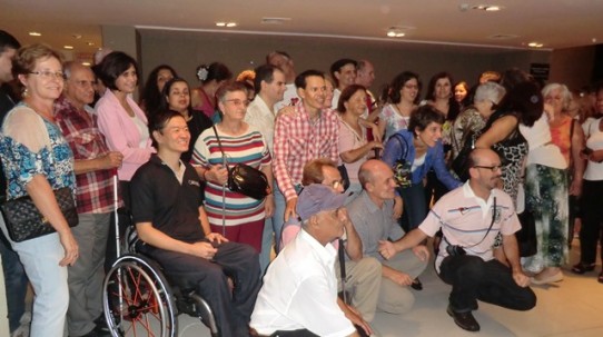 Fotografia colorida de várias pessoas com deficiência visual, posando para foto no hall do HSBC Brasil. No meio deles, Ricardo Shimosakai, do Turismo Adaptado.