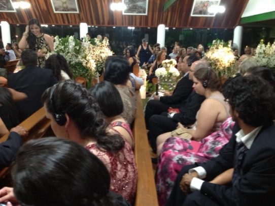 Fotografia colorida dos convidados sentados nos bancos da igreja decorada com arranjos de flores brancas, dentre eles várias pessoas com deficiência visual usando fones de ouvido.