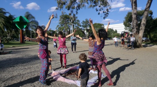 Fotografia colorida das bailarinas em pé, com os braços abertos para cima, em torno de um montinho de gravetos, cercados pelos tecidos esticados no chão, assemelhando-se a uma fogueira. Dois garotinhos aproximam-se com gravetos na mão e colocam na fogueira. Ao fundo, Lívia faz a audiodescrição segurando uma prancheta com o roteiro, Daniella Forchetti segura o pequeno Myguel, e embaixo da árvore,  estão Mari, Andréia, Cris e Talita Bretas.