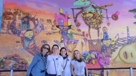 Fotografia colorida, em plano médio (da cintura para cima) de Lívia, Cristiana, Andréia e Rosângela, da VER COM PALAVRAS, todas sorridentes, em frente ao grande painel de arte urbana, com fundo lilás e personagens coloridos de pernas e braços finos, pintado por Os Gêmeos, na entrada do MAM.