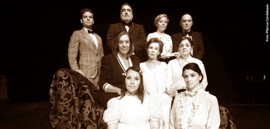 Fotografia em preto e branco de Dona Yayá, representada por Miriam Mehler, cercada por personagens que conviveram com ela até o fim de seus dias - três homens usando ternos e gravatas borboletas e três mulheres, com blusas fechadas, todos com fisionomias austeras; e por aqueles que estavam em sua memória, como as duas moças que estão em primeiro plano, sentadas no chão, com vestidos de mangas bufantes: Rosinha e D. Yayá moça, representada por Mariana Blanski. 