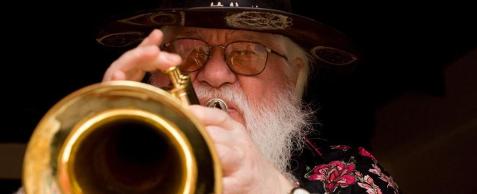 Fotografia colorida, em close, de Hermeto Pascoal tocando um trompete. Ele é um homem idoso de pele clara, com barba e cabelos brancos e longos. Tem os olhos pequenos e usa óculos. Usa camisa estampada e chapéu preto com rosetas na aba.