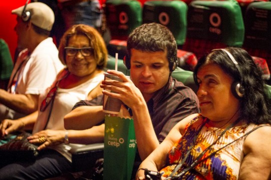 Descrição: Fotografia colorida, em primeiro plano (do peito para cima), de Diniz Cândido, que segura um saco de pipocas e refrigerante, ao lado de Margarida. Os dois usam fones de ouvido. José Vicente de Paula e Marta Gallo estão na mesma fileira, à esquerda. (foto Raoni Reis)