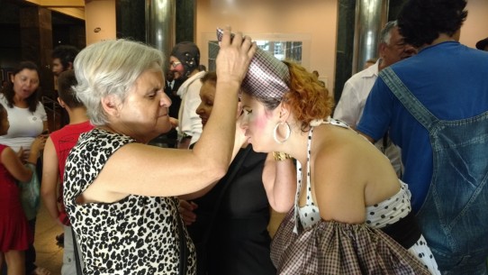 Fotografia colorida de D. Lucinha tocando o grande laço prata que está nos cabelos de Maria Vilhosa. D. Lucinha é uma senhora de cabelos brancos curtos, penteados de lado; usa vestido bege com estampas pretas de pele de onça. Maria Vilhosa é uma mulher de pele clara, cabelos louros, curtos e cacheados; usa um vestido frente única, branco com bolinhas pretas, com saia rodada na altura dos joelhos e um cinto preto. 