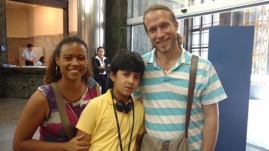 Fotografia colorida em plano médio (da cintura para cima), de Lukas, um garoto moreno, de cabelos pretos com franja e camiseta amarela com seus pais, Melina e Jonathan, no hall da Cultural, aguardando a apresentação do espetáculo infanto-juvenil: ERA UMA ERA.