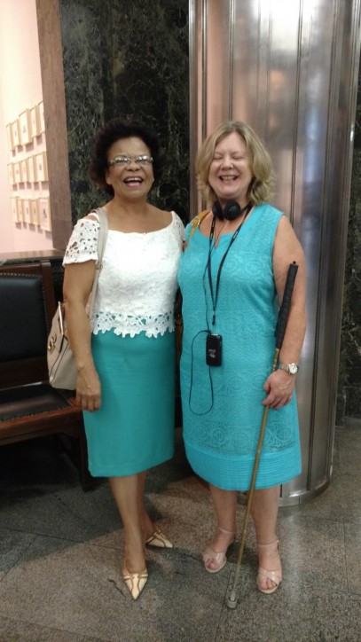 Fotografia colorida de corpo inteiro, de Maria Regina Lopes ao lado de sua amiga Rose, as duas sorridentes posando para foto, no hall da Caixa Cultural, aguardando a apresentação do espetáculo Poema Suspenso para uma Cidade em Queda. Regina é uma mulher loura de cabelos na altura dos ombros, olhos verdes. Usa vestido sem mangas azul piscina. Rose tem os cabelos castanhos curtos, usa óculos, blusa branca de renda e saia azul piscina.