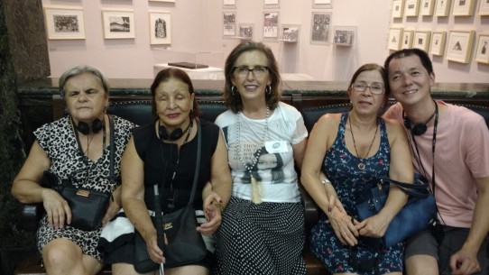 Fotografia colorida de Lívia sentada em um banco de couro, no meio dos amigos: D. Lucinha, Marilene de Paula, Maria de Lourdes e Edson Kadihara, no hall da Caixa Cultural, tendo ao fundo muitos quadros pequenos na parede.