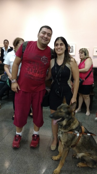 Fotografia colorida de Guilherme Rubino e Marina Guimarães, os dois sorridentes, abraçados, posando para foto, aguardando a apresentação do espetáculo Poema Suspenso para uma Cidade em Queda.  Ao lado de Marina, o cão-guia Duke. Marina é uma jovem de pele clara, cabelos castanhos lisos e longos, repartidos ao meio e sorriso largo. Ela usa um minivestido preto e bege sem mangas. Guilherme tem cabelos bem curtos, o rosto redondo e usa camiseta e bermuda cor de vinho.