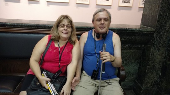 Fotografia colorida Milene Orofisi e Markiano Charan sentados em um banco de couro, no hall da Caixa Cultural, aguardando para assistir ao espetáculo Poema Suspenso para uma Cidade em Queda. Os dois sorridentes com camisetas regatas e bermudas, fones no pescoço e receptores pendurados no peito, seguram suas bengalas.