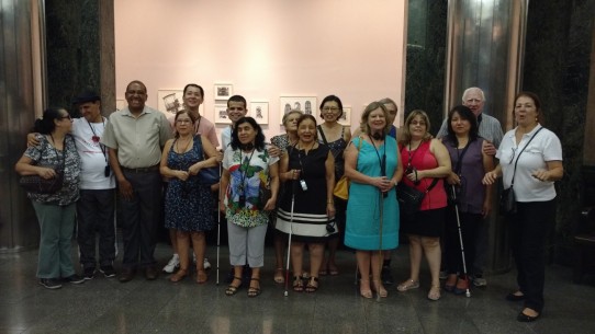 Fotografia colorida de um grupo de pessoas com e sem deficiência visual, no hall da Caixa Cultural, aguardando para assistir ao espetáculo Poema Suspenso para uma Cidade em Queda. Da esquerda para direita: D. Eva, José Vicente de Paula, Luciano, Edson Kadihara, Maria de Lourdes, Diniz Cândido, Margarida, D. Lucinha, Marilene de Paula, Amélia Hashimoto, Markiano Charan, Milene Orofisi, Regina Lopes, Marco Bertolli, Kátia Chimabokuro e Fátima Angelo.