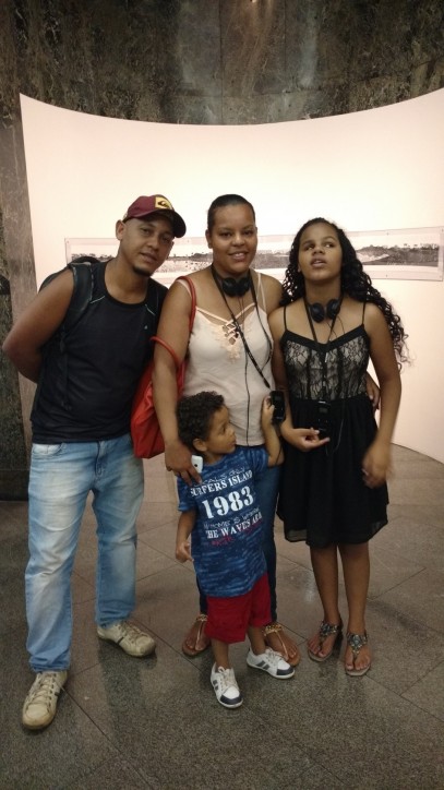 Fotografia colorida de corpo inteiro, de Ingrid, uma garota morena de longos cabelos crespos, com seus pais, Josefa e Álvaro, e seu irmãozinho, no hall da Caixa Cultural, aguardando a apresentação do espetáculo infanto-juvenil: ERA UMA ERA.