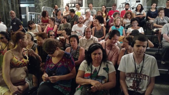 Fotografia colorida da plateia do Teatro da Caixa Cultural, com muitas pessoas com deficiência visual com fones de ouvido e receptores, aguardando o início do espetáculo Poema Suspenso para uma Cidade em Queda. Da esquerda para direita, na primeira fileira: Iris, Eliana Cynthia, Margarida e Diniz Cândido; na segunda fileira: Rosenir, D. Lucinha, Marilene de Paula, Maria Eva, Edson Kadihara e Maria de Lourdes: na terceira fileira: D. Eva, Amélia Hashimoto, Luciano e Sr. Aguinaldo; na quarta: Regina Lopes e Rose.