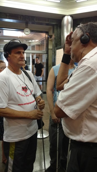 Fotografia colorida em plano americano (dos joelhos para cima) de José Vicente de Paula, da ONG Amigos Prá Valer, conversando com Sr. Aguinaldo Batista, no hall da Caixa Cultural, aguardando para assistir ao espetáculo infanto-juvenil: Era uma Era. 