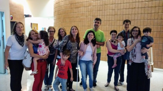 Fotografia colorida de um grupo de pais, mães e uma avó com deficiência visual acompanhados dos filhos e neta com os audiodescritores da VER COM PALAVRAS, posando para foto, no hall do Teatro Sérgio Cardoso, logo após o espetáculo O Gigante ao Vivo do Grupo Tiquequê. Da esquerda para direita, Fátima Angelo, Margarete Jardim com a neta Manuela no colo, Sônia Ferreira, Marina Caruso, Clarissa Sanches com a mão no ombro de Lourenço, Maria Rita Paiva, Cleber Souza, Priscila Teixeira com sua filhinha Julia e o marido Carlos Massao, e Lívia com Raul, filho de Maria Rita e Cleber, no colo.