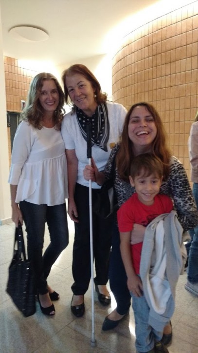 Fotografia colorida de Clarissa Sanches sorridente, abaixada e apoiada na bengala, segurando o pequeno Lourenço com a mão no peito dele. Clarissa está com um casaco de moleton pendurado no braço. Atrás deles, Marina Caruso e Fátima Angelo. Marina é loura, de cabelos longos e anelados, usa bata branca de mangas ¾, calça jeans e segura uma bolsa preta. Fátima tem os cabelos castanhos na altura dos ombros, os olhos pequenos, usa blusa branca com echarpe preta de poás brancos e calça preta.