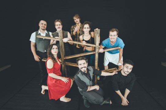 Fotografia colorida dos oito artistas circenses do grupo Cia Los Circo Los, seis homens e duas mulheres, todos sorridentes, ao redor de uma estrutura de madeira com canaletas transversais. Cinco artistas estão em pé e três estão abaixados.
