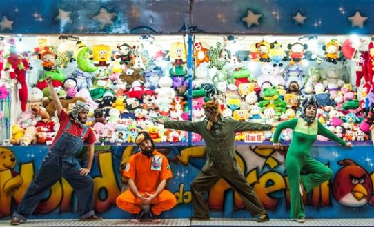 Fotografia colorida dos quatro integrantes da banda Fera Neném, na frente de uma barraca repleta de bichinhos de pelúcia de um parque de diversões. Da esquerda para a direita: Gustavo Cabelo, Pedro Gongom, Peri Pane e Lia Biserra. Gustavo está em pé com uma mão para o alto e a outra no joelho; veste camiseta vermelha, jardineira jeans e uma touca gorro de tigre siberiano. Pedro está agachado; veste macacão laranja de mangas curtas e uma touca gorro de macaco. Peri está em pé com os braços abertos; veste macacão verde de mangas compridas e uma touca gorro de vaquinha. Lia está em pé com as mãos apoiadas na barraca; veste macacão verde de mangas compridas e uma touca gorro de elefante.