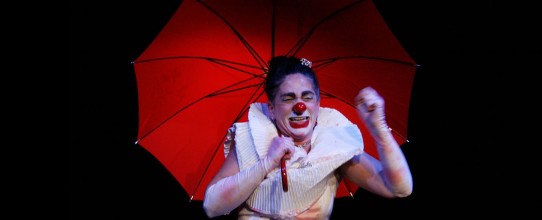 Fotografia colorida na horizontal em plano médio sobre fundo escuro da palhaça Mafalda Mafalda, interpretada por Andréa Macera, segurando um guarda chuva vermelho, com a boca entreaberta e os olhos contraídos, parecendo chorar. Ela é uma mulher de pele clara, cabelos castanhos presos em um coque alto, sobrancelhas escuras e espessas. O rosto está pintado de branco, o lábio inferior de vermelho e o nariz é uma bolinha vermelha. Veste uma blusa branca sem mangas com uma grande gola bufante. Usa luvas três quartos manchadas.