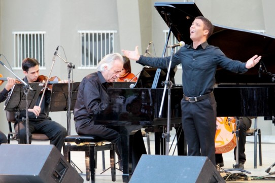 Fotografia colorida do tenor Jean William cantando com os braços abertos ao lado do Maestro João Carlos Martins que está sentado ao piano. Jean é um jovem negro, de cabelos castanhos curtos. O maestro tem a pele clara, os cabelos brancos e cheios. Os dois usam camisas de mangas longas e calças pretas. Ao fundo, três músicos, dois homens e uma mulher, tocam violinos.