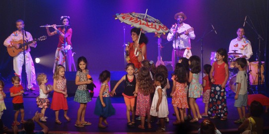 Fotografia colorida com fundo azulado dos cinco componentes do grupo CantaVento (três homens e duas mulheres) no palco, tocando seus instrumentos e cantando, cercado de muitas crianças. Carol Ladeira está à frente do grupo, segurando um microfone e uma sombrinha aberta.