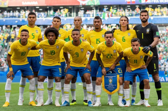 Fotografia colorida do time de futebol do Brasil, com dez jogadores de camisas amarelas com golas verdes, shorts azuis e meias brancas até os joelhos e um jogador, o goleiro, de camisa, shorts, luvas e meias pretas. Os jogadores estão organizados em duas fileiras. Na fileira de trás, da esquerda para direita estão: Casemiro, Thiago Silva, Miranda, Paulinho, Filipe Luis e o goleiro Alisson. Na frente, ligeiramente abaixados, estão: Neymar, William, Gabriel Jesus, Fagner que segura a flâmula do time e Philippe Coutinho. Ao fundo, a imagem desfocada da arquibancada lotada do estádio.